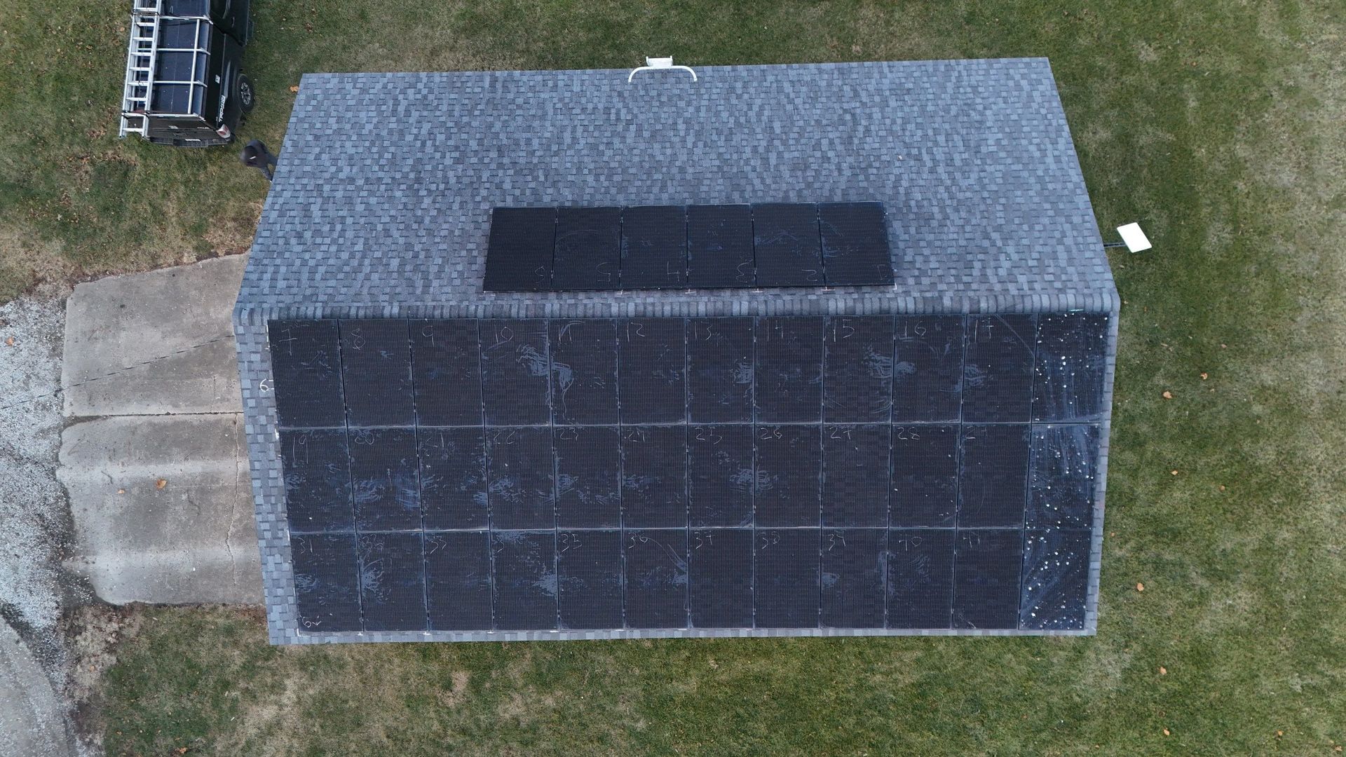 Overhead view of a dark gray rooftop with solar panels; small shed in grassy area.