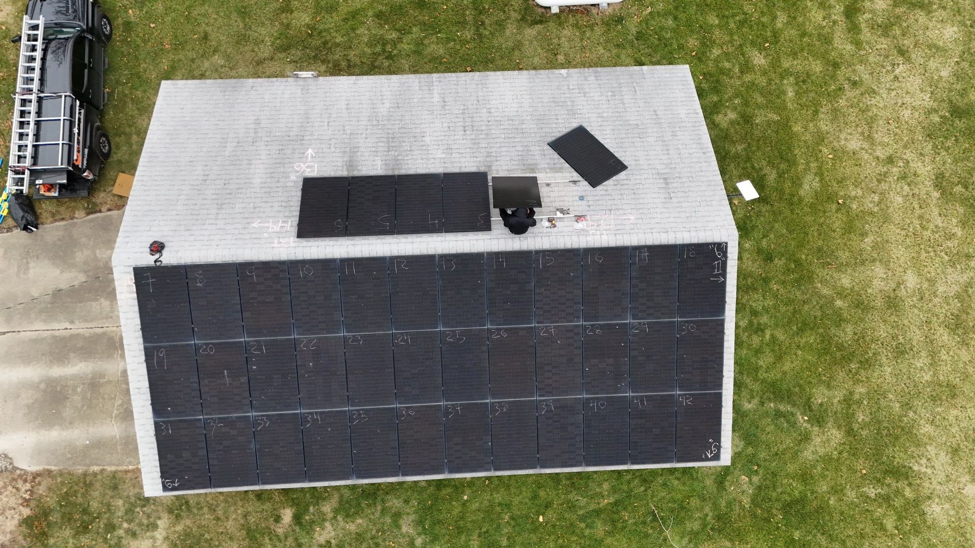 A roof partially covered with new black shingles, with a truck and ladder nearby, and grass surrounding the house.