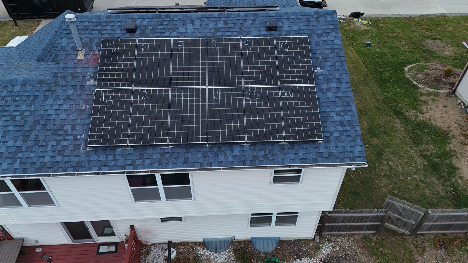 Solar panels on a two-story house with a blue roof, near a grassy area and wooden fence.