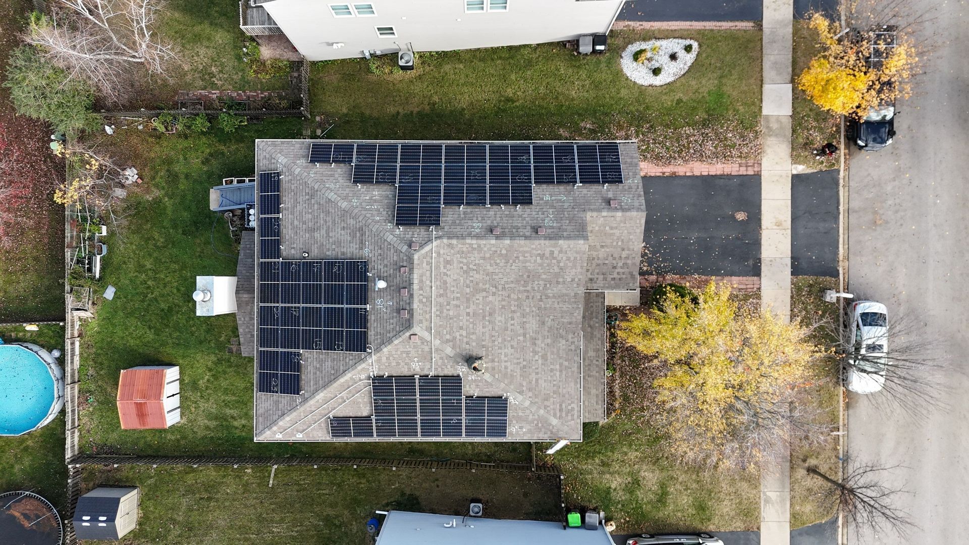 Overhead view of a house with solar panels on the roof, surrounded by green grass and autumn trees.