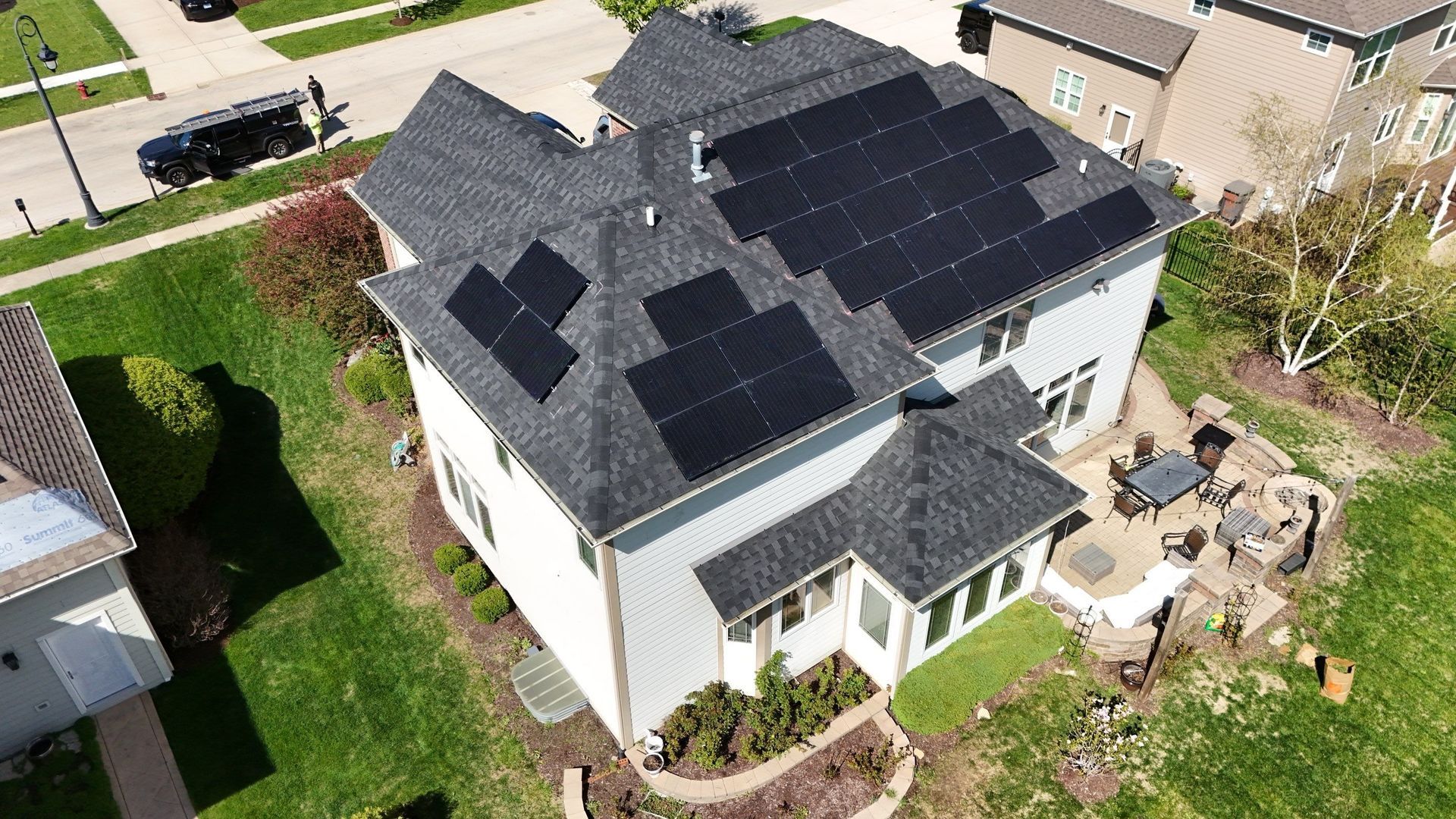 An aerial view of a two-story house with solar panels on the roof. Green lawn and a patio are also visible.