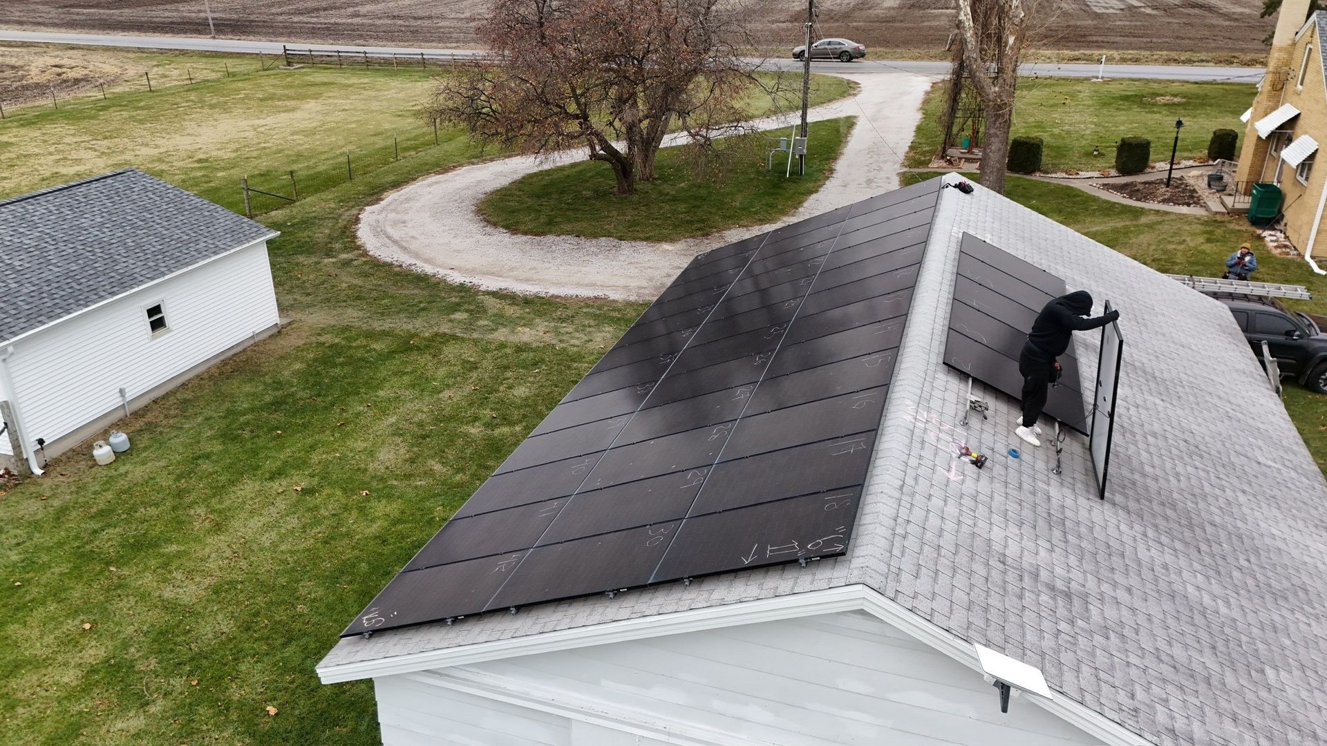 Person installing solar panels on a gray-roofed house.  Green lawn surrounds the house and a white outbuilding is nearby.