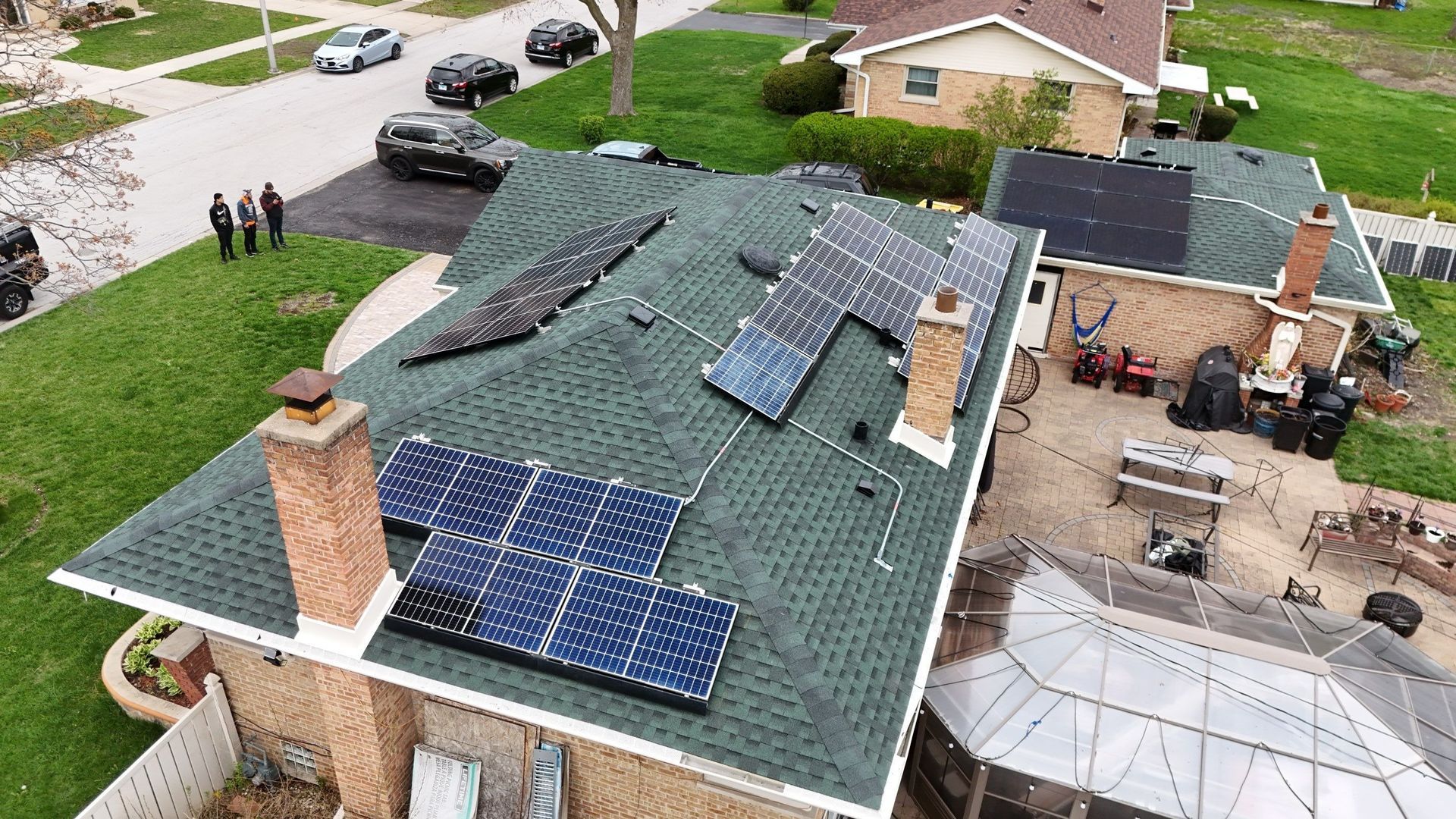 Rooftop view of house with solar panels installed on a green shingle roof. Cars and people in the background.