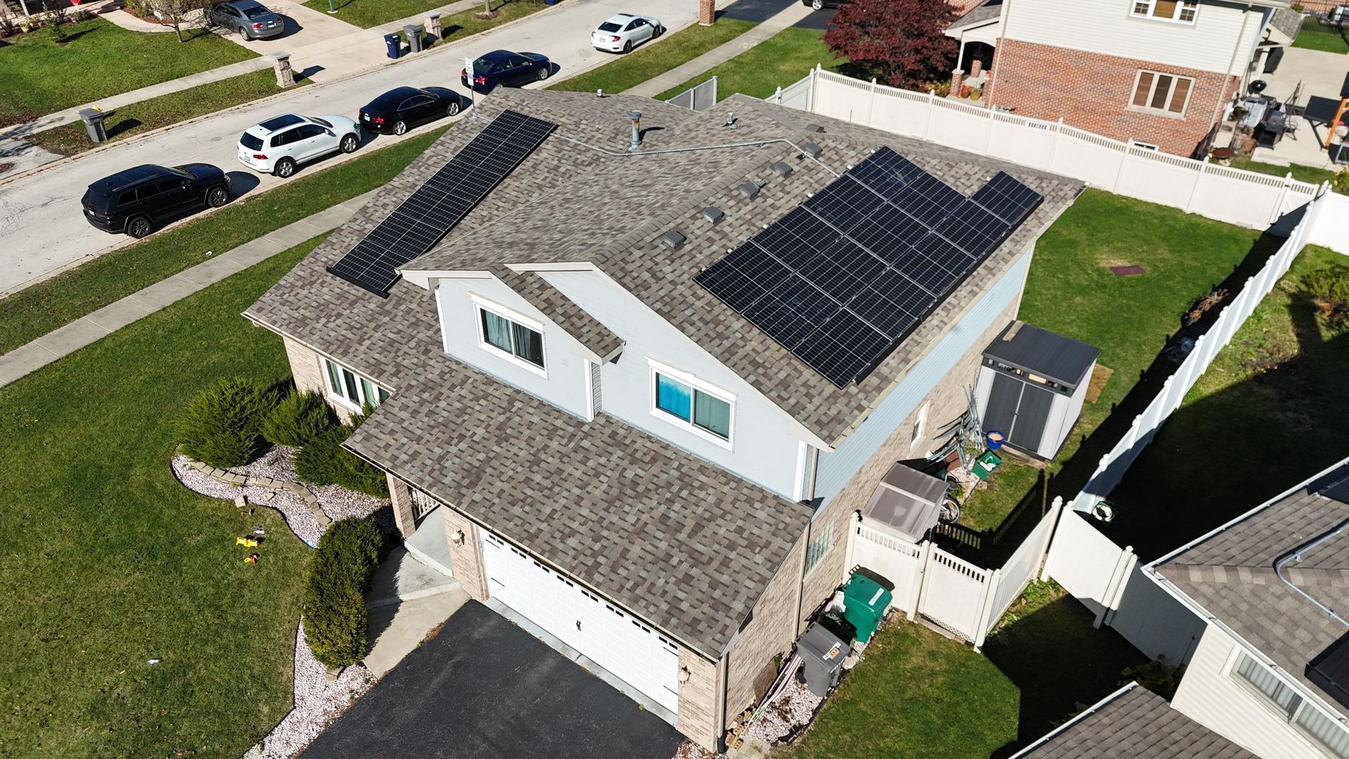 Aerial view of a house with solar panels on the roof, green lawn, and street with cars.