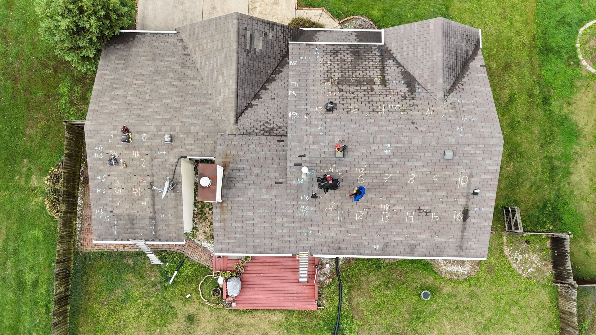 Overhead view of a house with a weathered roof, surrounded by green grass. Two figures are on the roof.