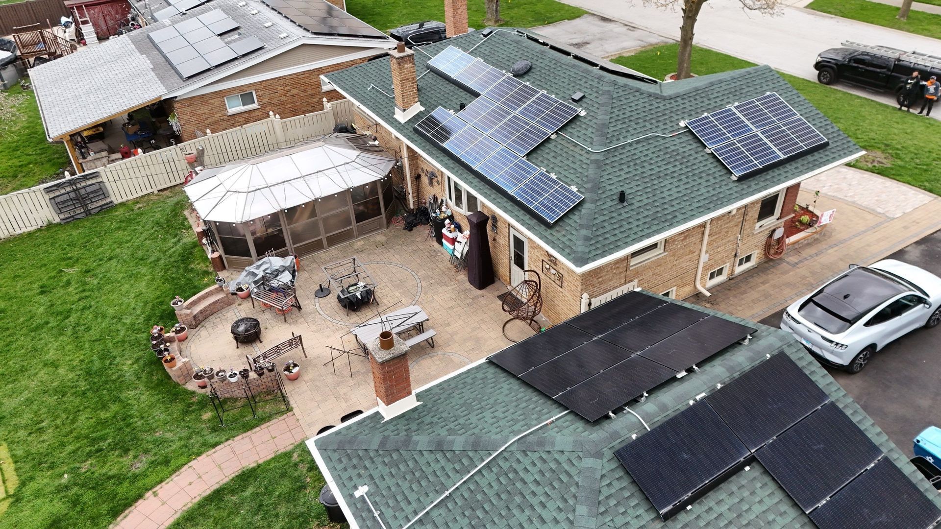 An aerial view shows homes with solar panels on green roofs, including a house with a patio and an EV in the driveway.
