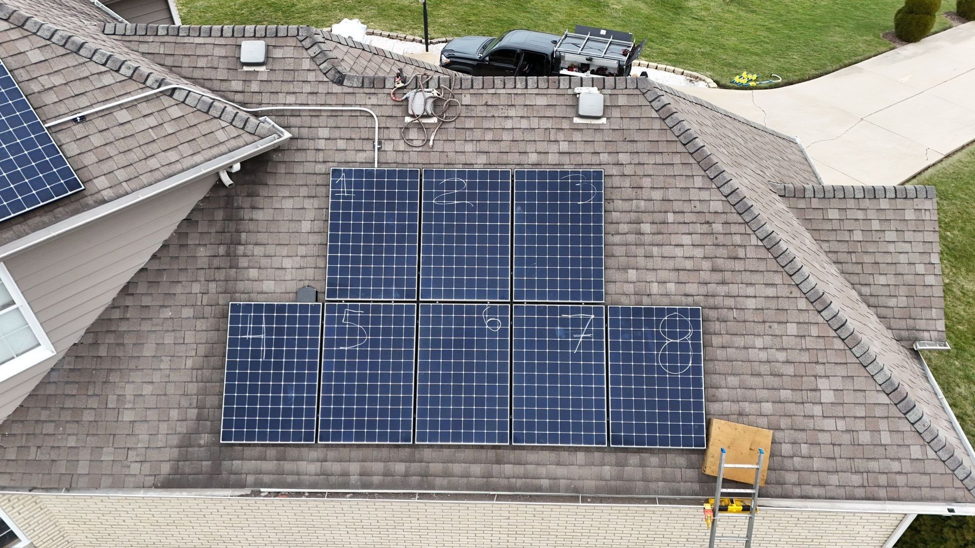 Solar panels on a brown shingled roof. A ladder and vehicle are also visible.
