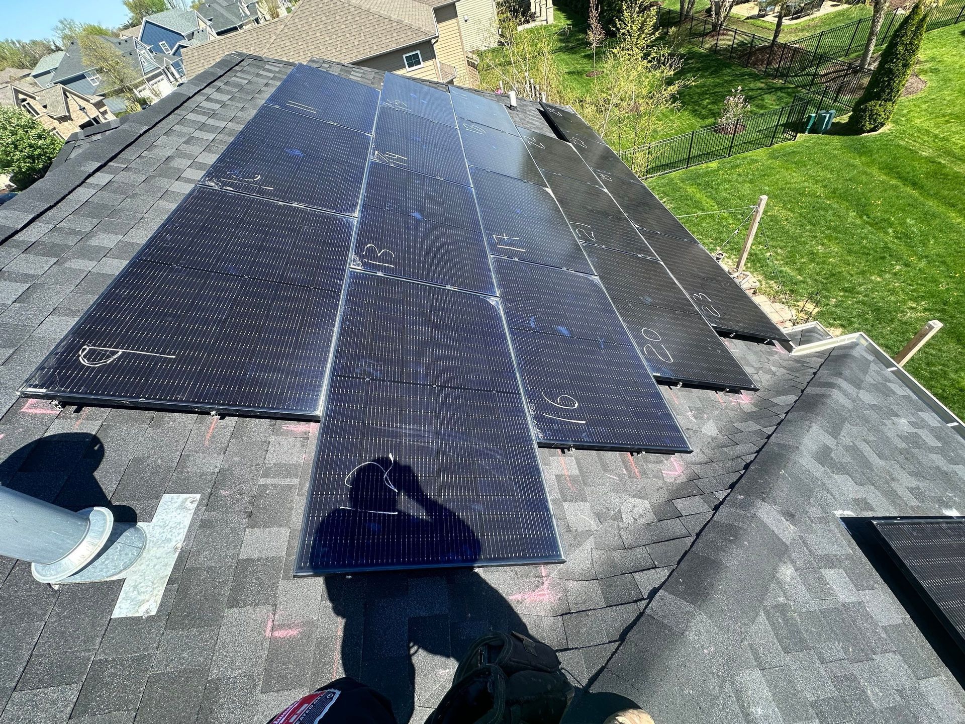 Solar panels installed on a dark shingled roof, reflecting the sky, with a grassy yard in the background.