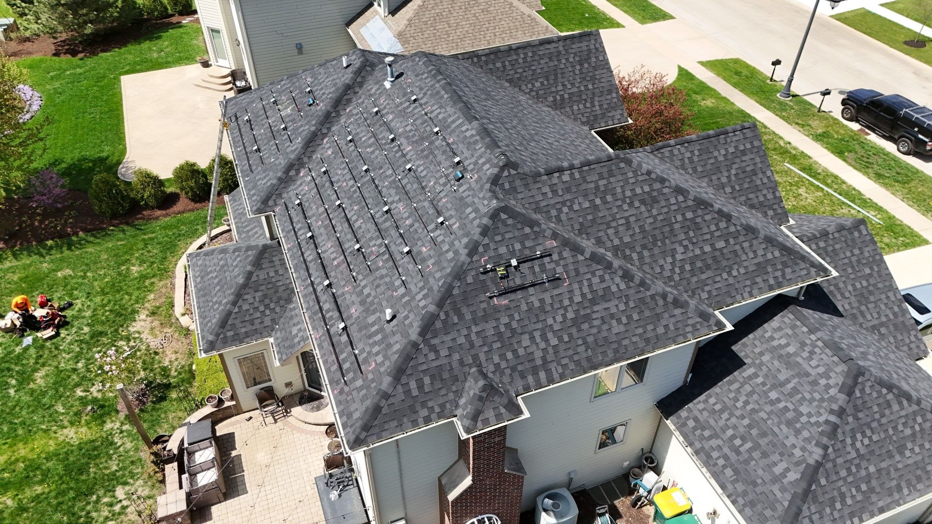 Overhead view of a large house with a dark gray shingled roof on a sunny day; green lawn surrounds the house.