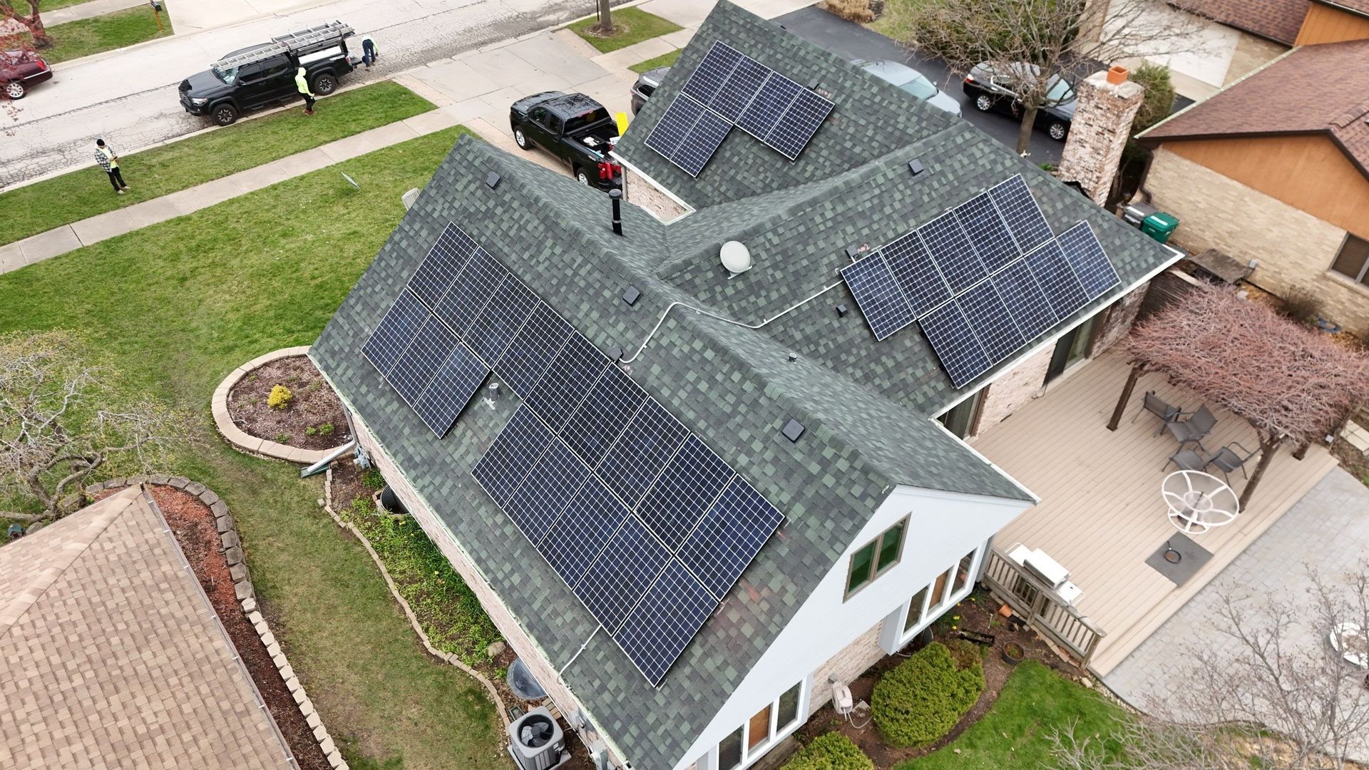 Aerial view of a house with solar panels on its roof. It's surrounded by green grass and other houses.