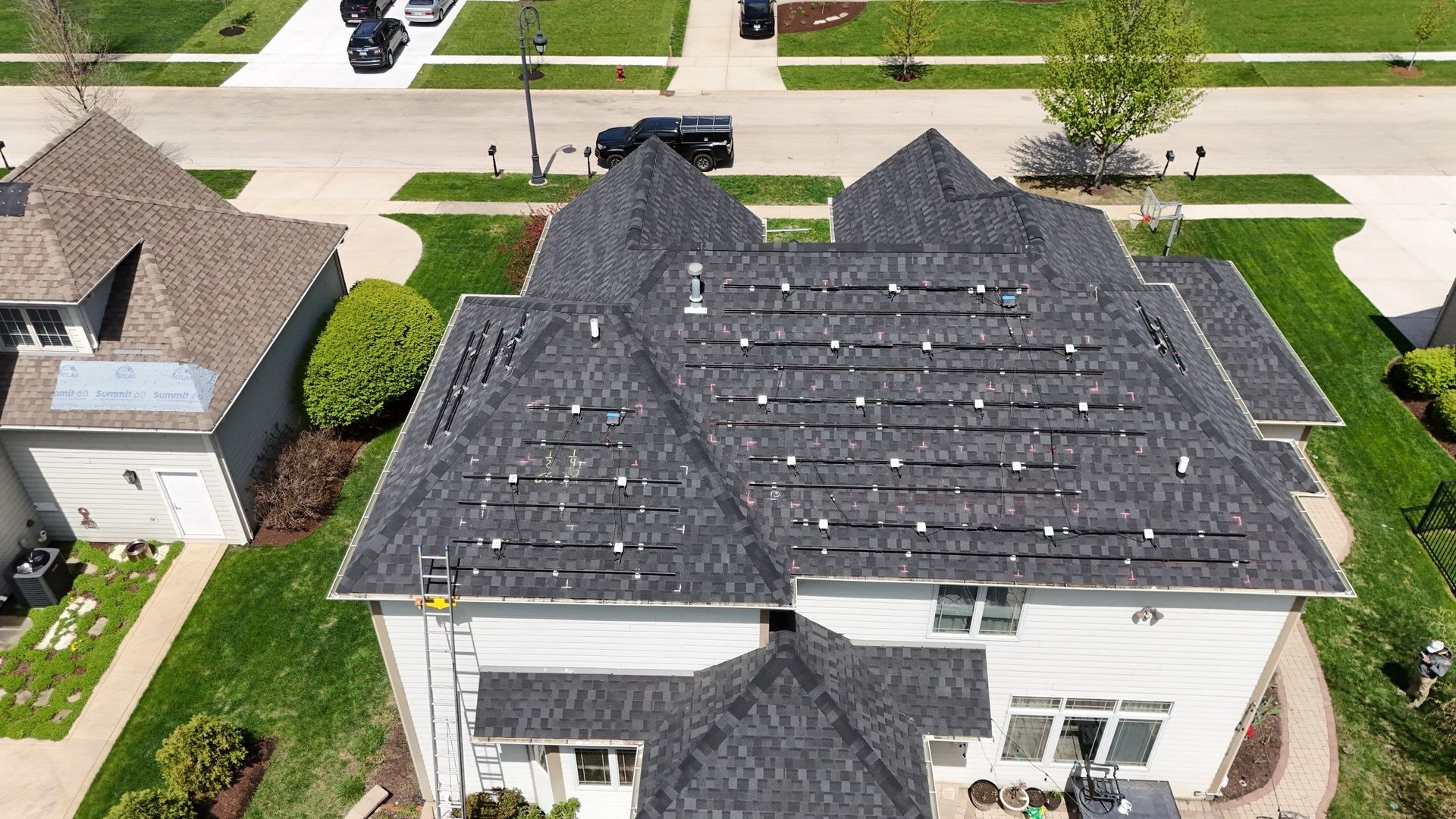 Aerial view of a house with dark shingles and installed solar panels. Green lawn surrounds it in a suburban neighborhood.