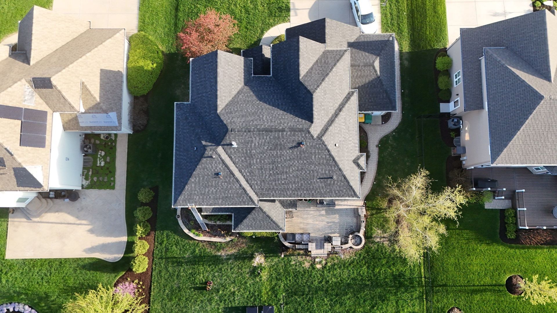 Overhead view of a multi-roofed house with a deck, surrounded by green lawns and neighboring homes.