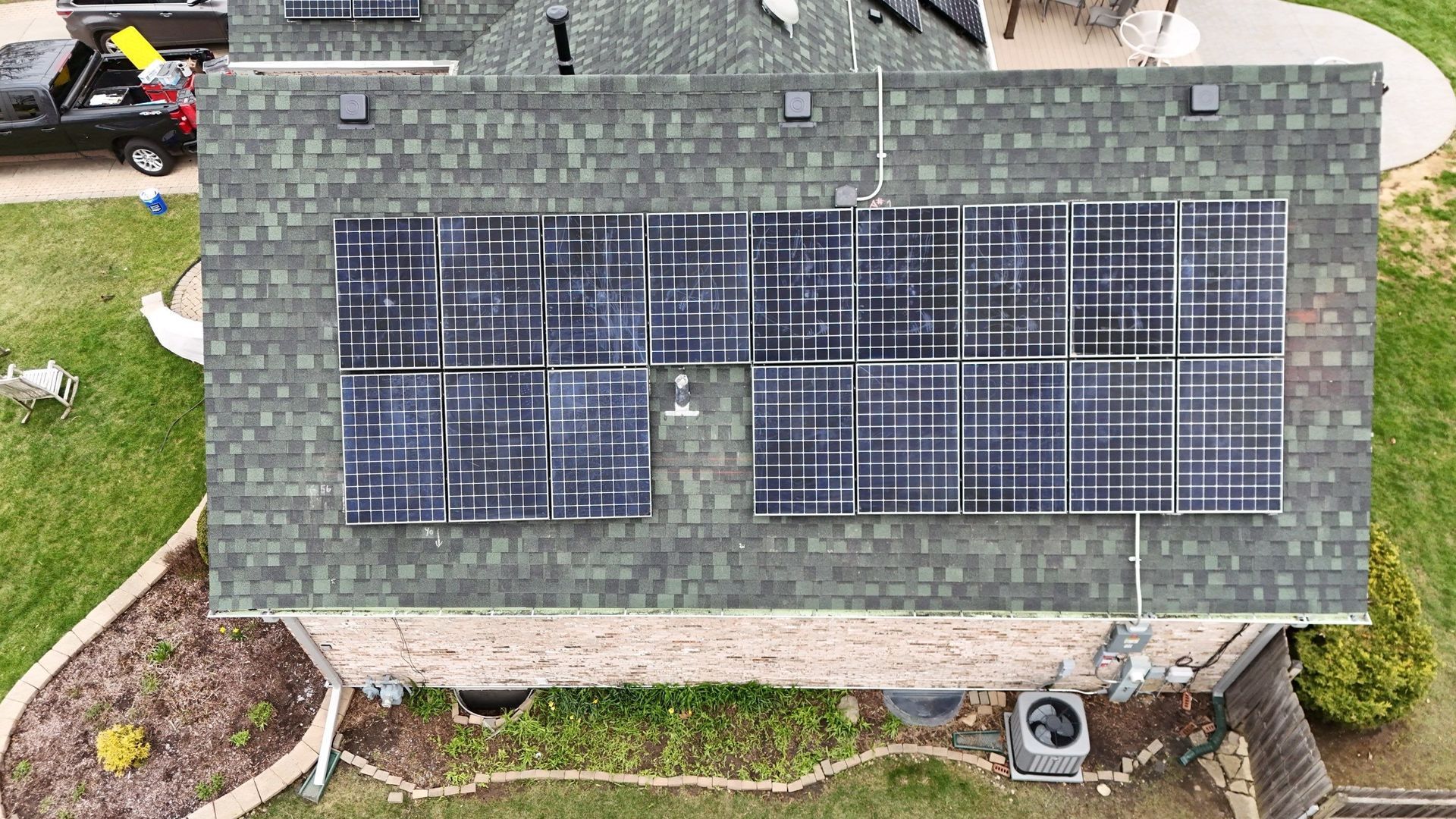 Solar panels installed on a green shingled roof. A truck, green grass, and a brick house are also visible.