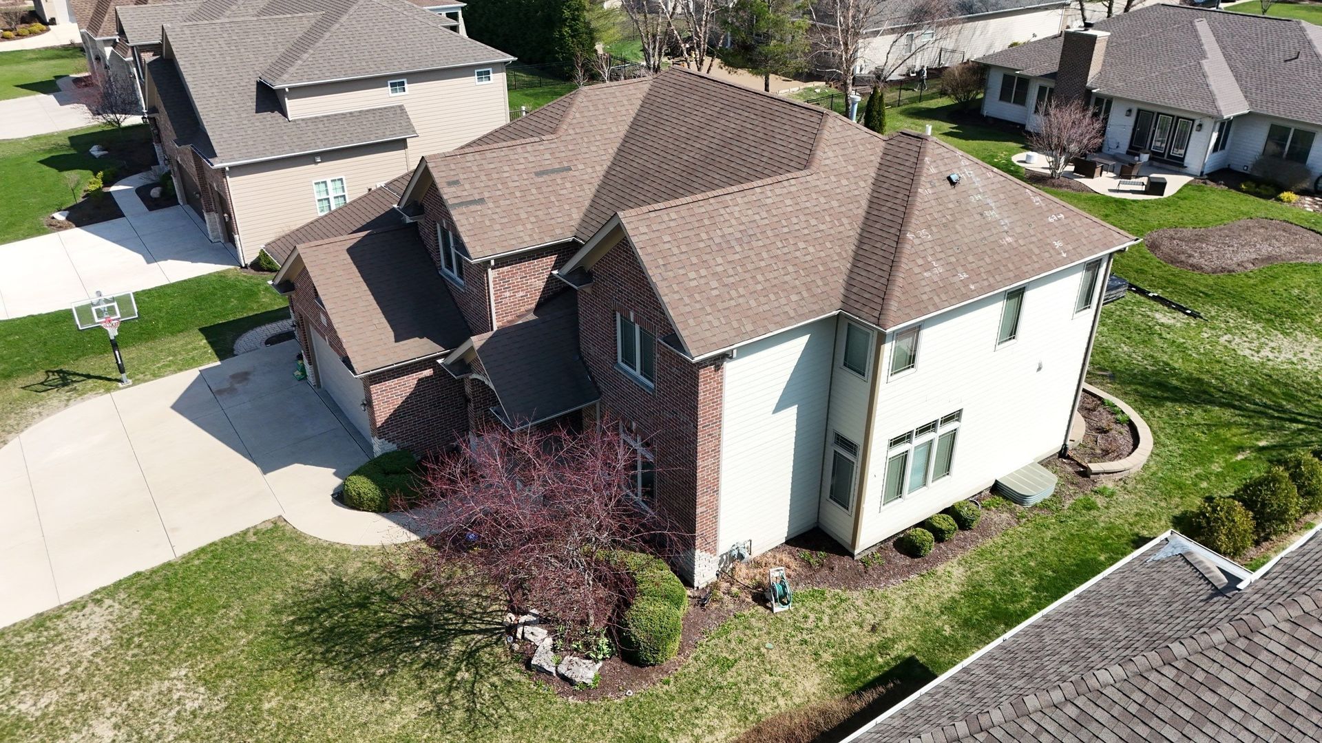 Two-story house with brown roof, brick and light-colored siding, surrounded by green grass and other houses.
