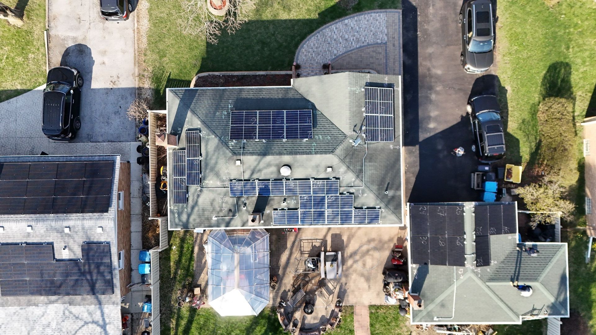 Overhead view of a house with solar panels on the roof, parked cars in the driveways, and surrounding houses.