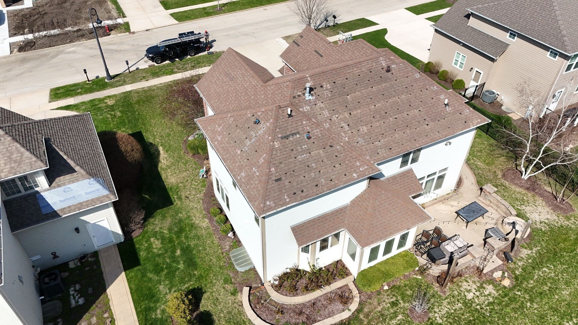 Aerial view of a two-story white house with a brown roof and small patio. Green grass and neighboring homes are visible.