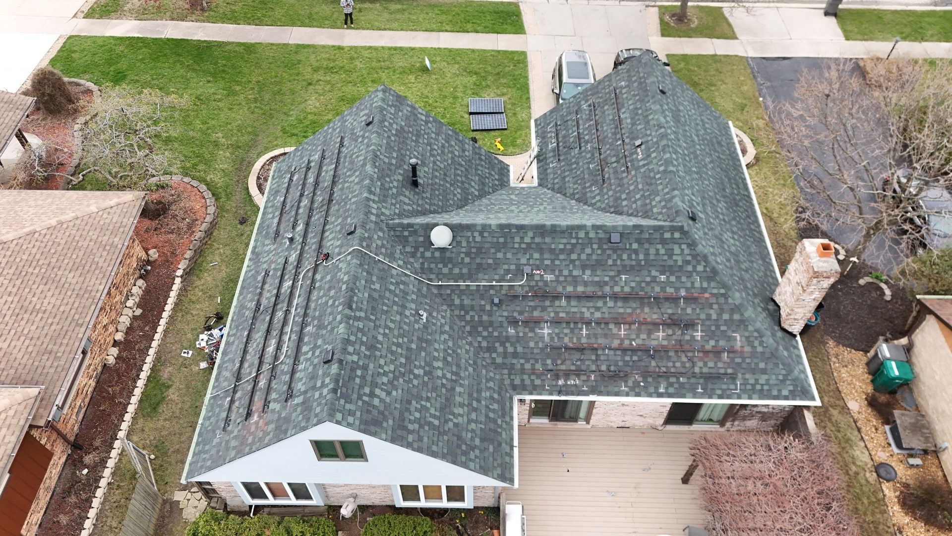 Overhead view of a house with green roof, surrounded by grass, a driveway, and a garden.