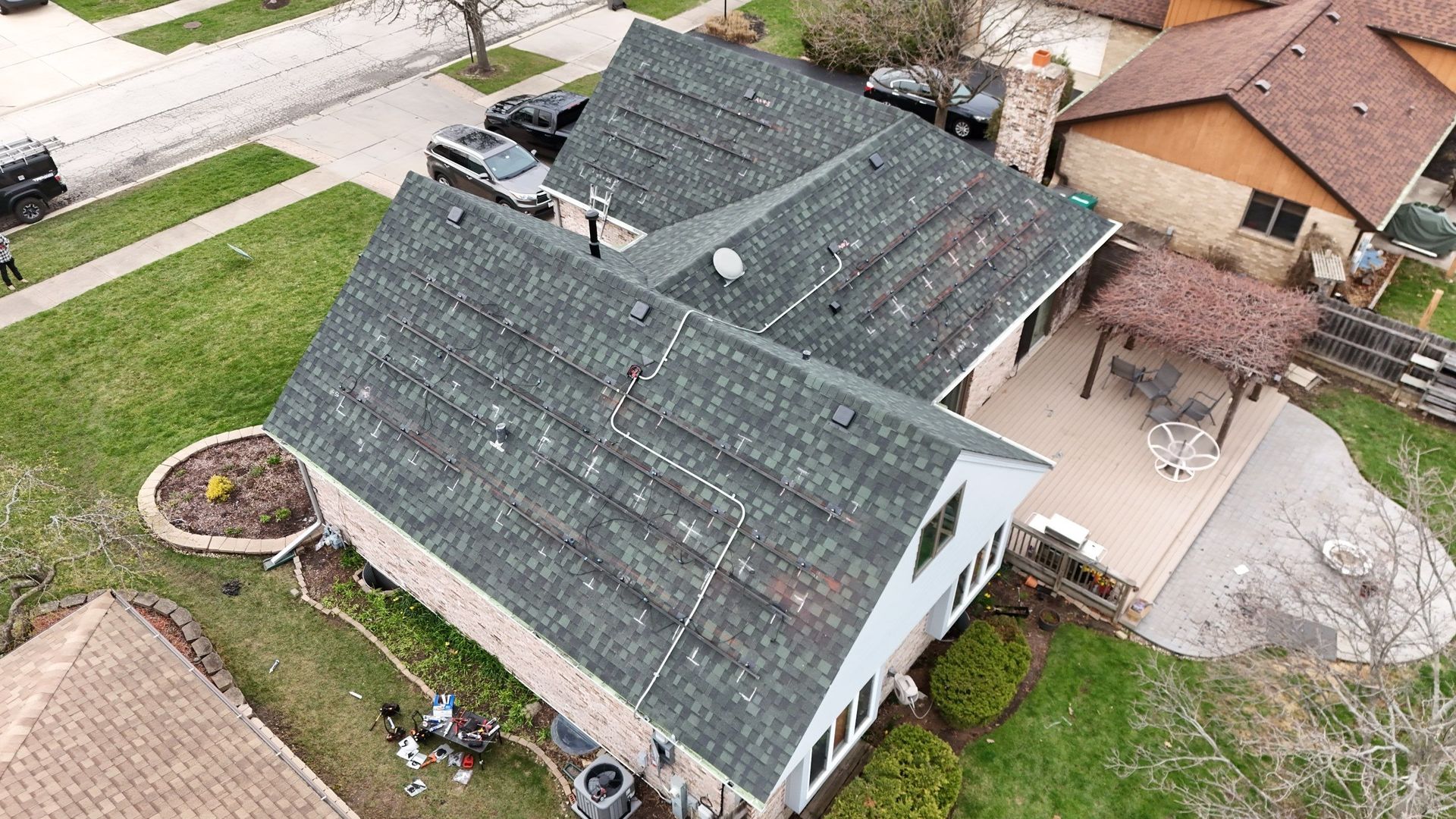 Aerial view of a house with dark green shingled roof and a backyard patio; trees and grass.