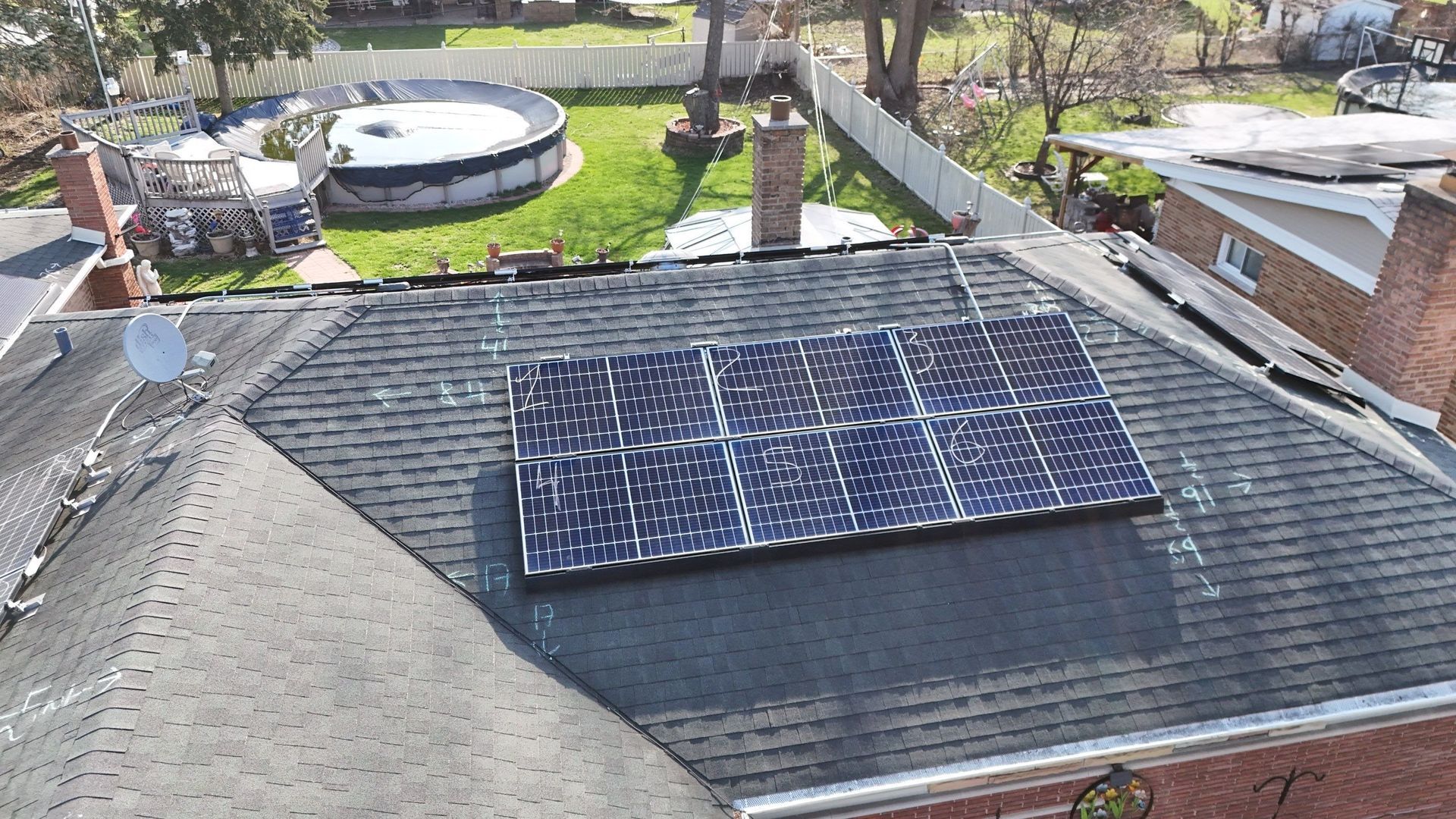 Solar panels on a dark shingled roof of a brick house, with a backyard including a pool and white fence.