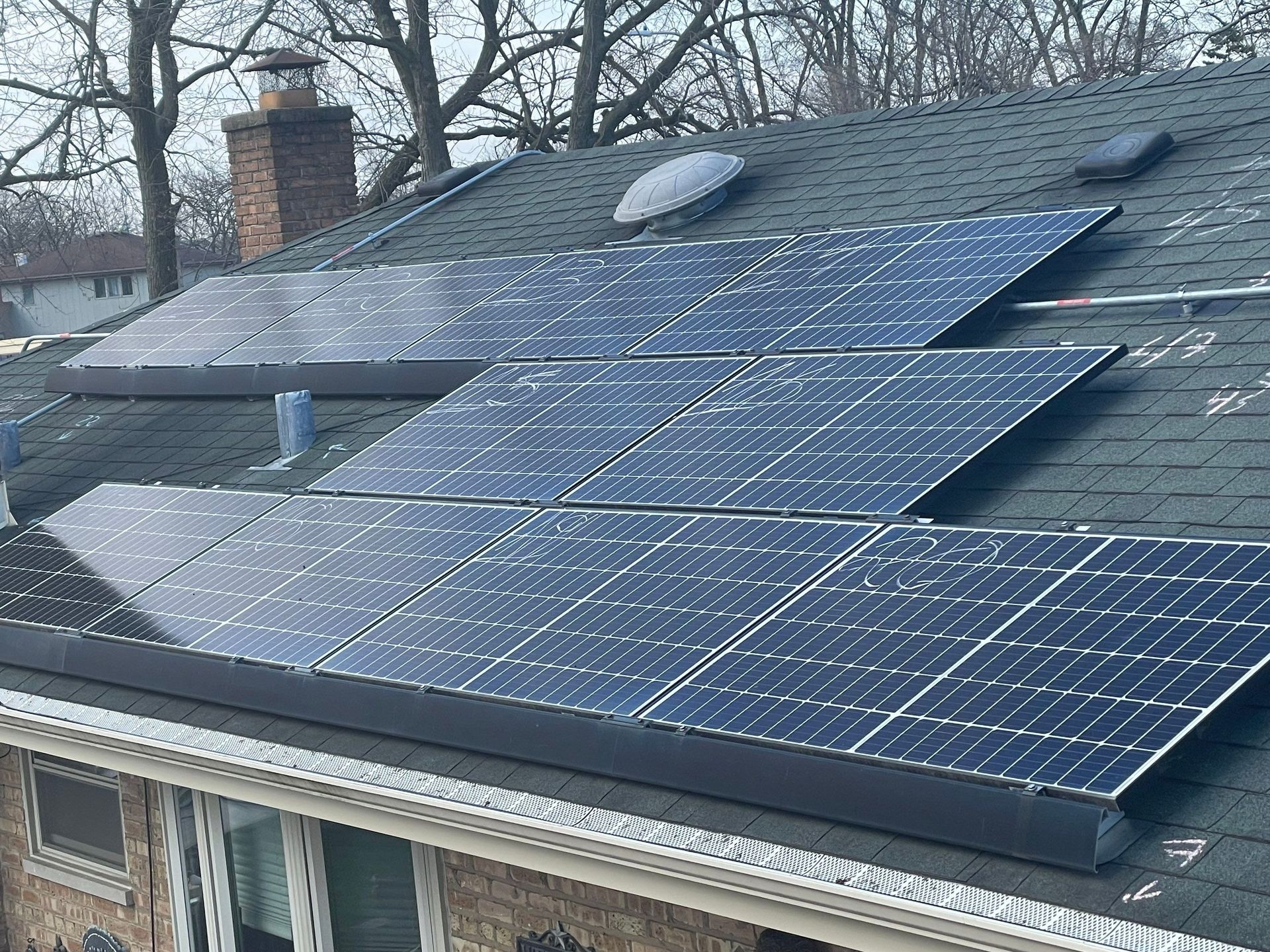 Solar panels installed on a dark-shingled roof of a residential house with bare trees in the background.