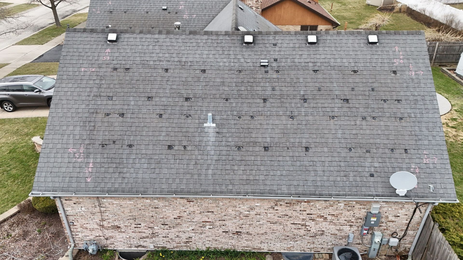 Overhead view of a gray asphalt shingle roof on a brick house; visible vents, satellite dish, and air conditioning unit.