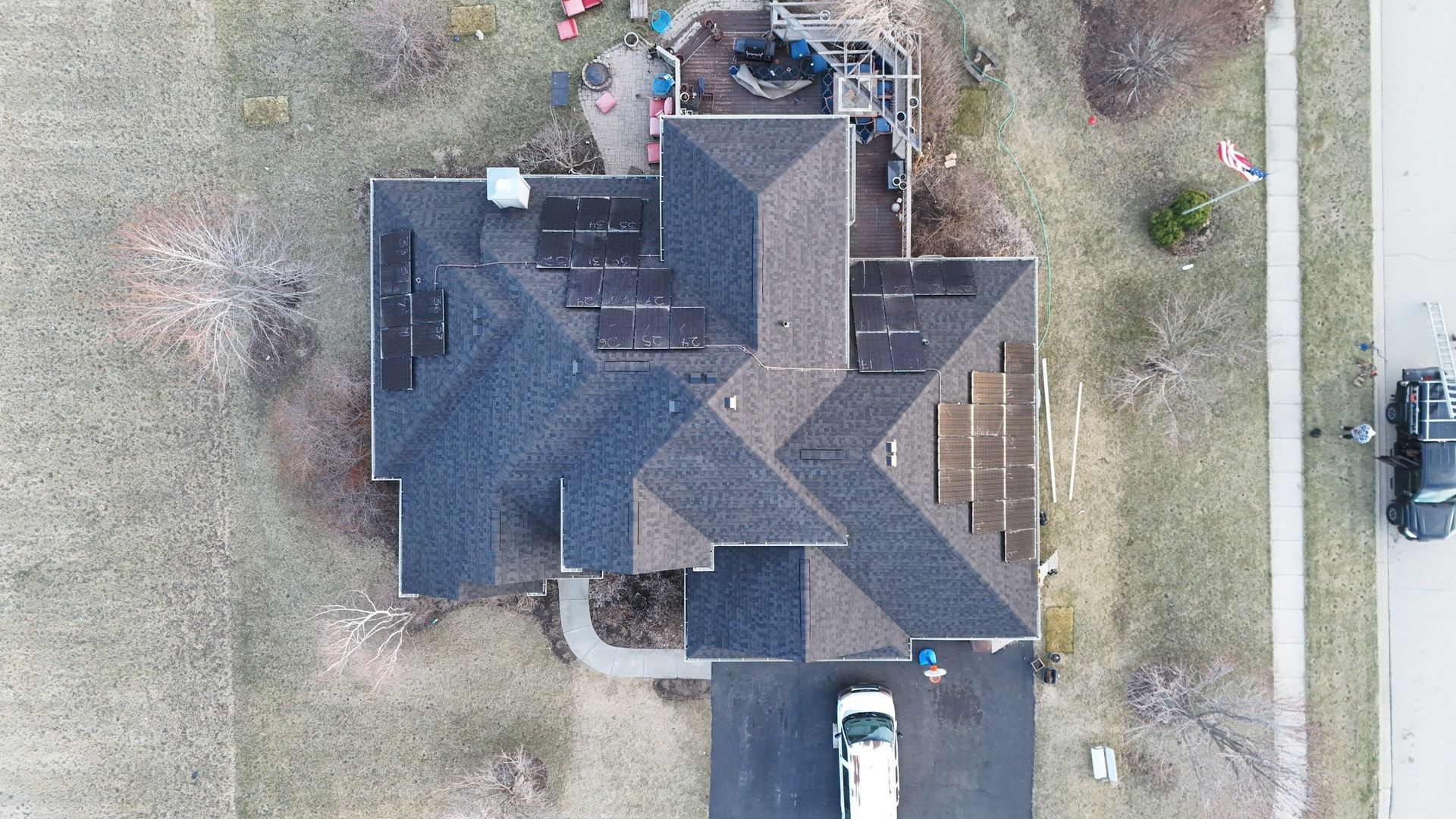 Overhead view of a house with dark gray shingles, solar panels, surrounded by grass, with a driveway and street visible.