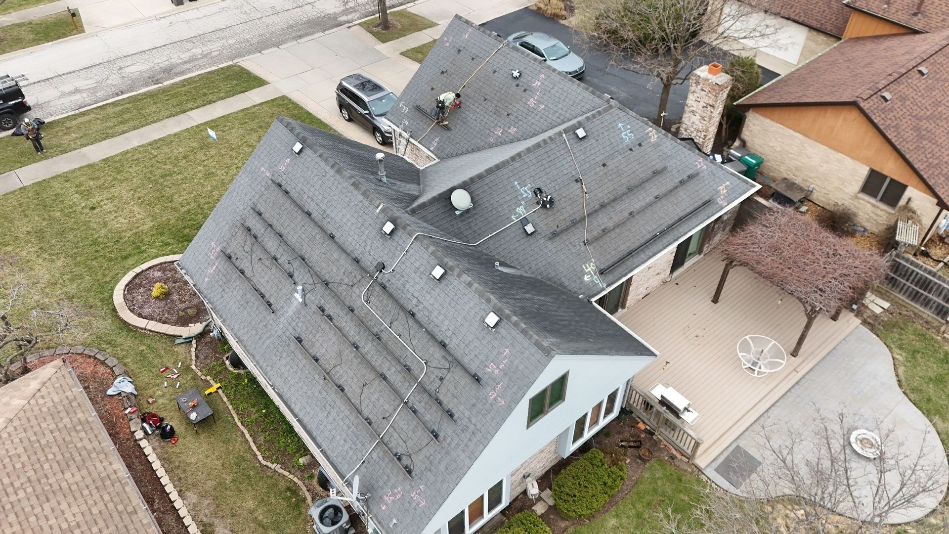 Aerial view of a house with solar panels on the roof. Gray asphalt shingles, cars in driveway, and a wooden deck are visible.