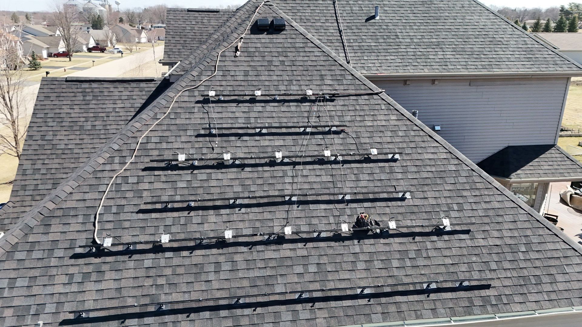 Rooftop with rows of solar panels installed on dark asphalt shingles. Houses are visible in the background.