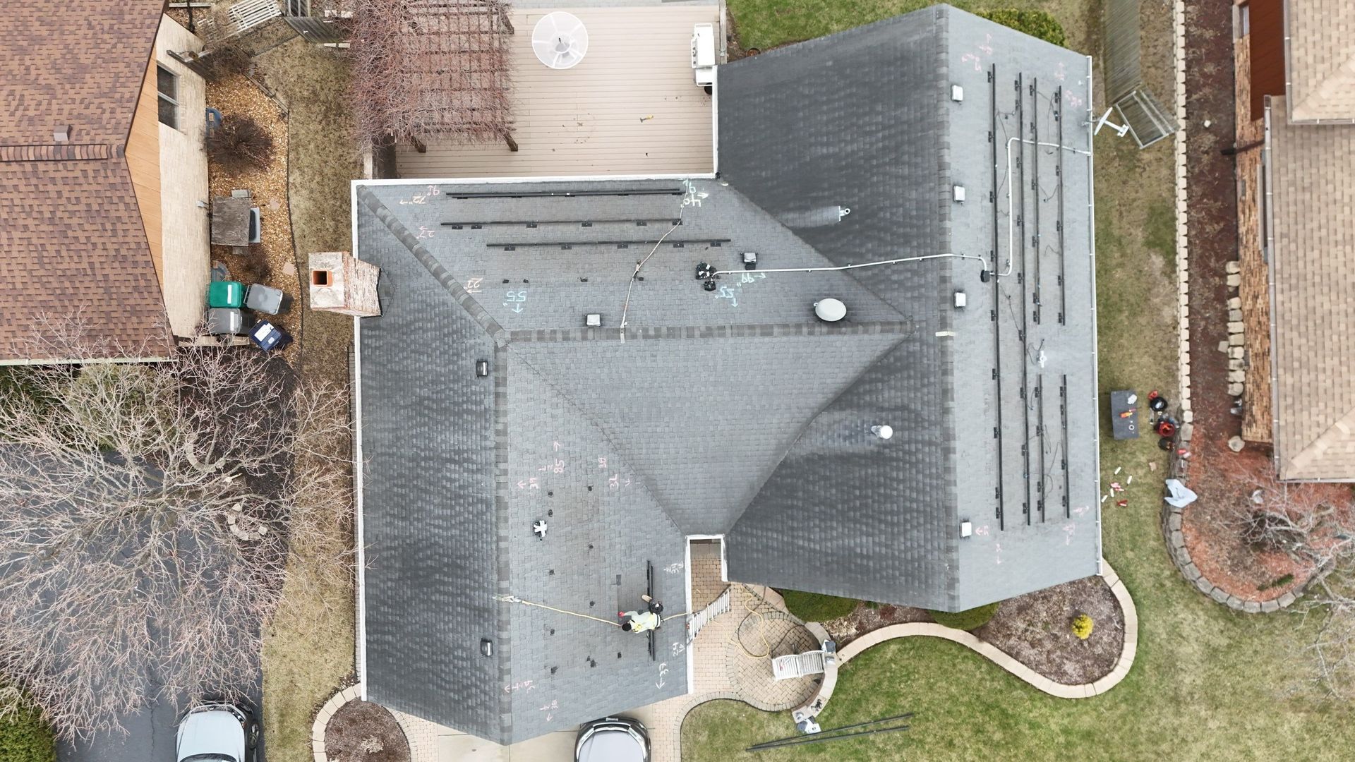Aerial view of a house with gray shingles, some solar panels, and surrounding yard with greenery.