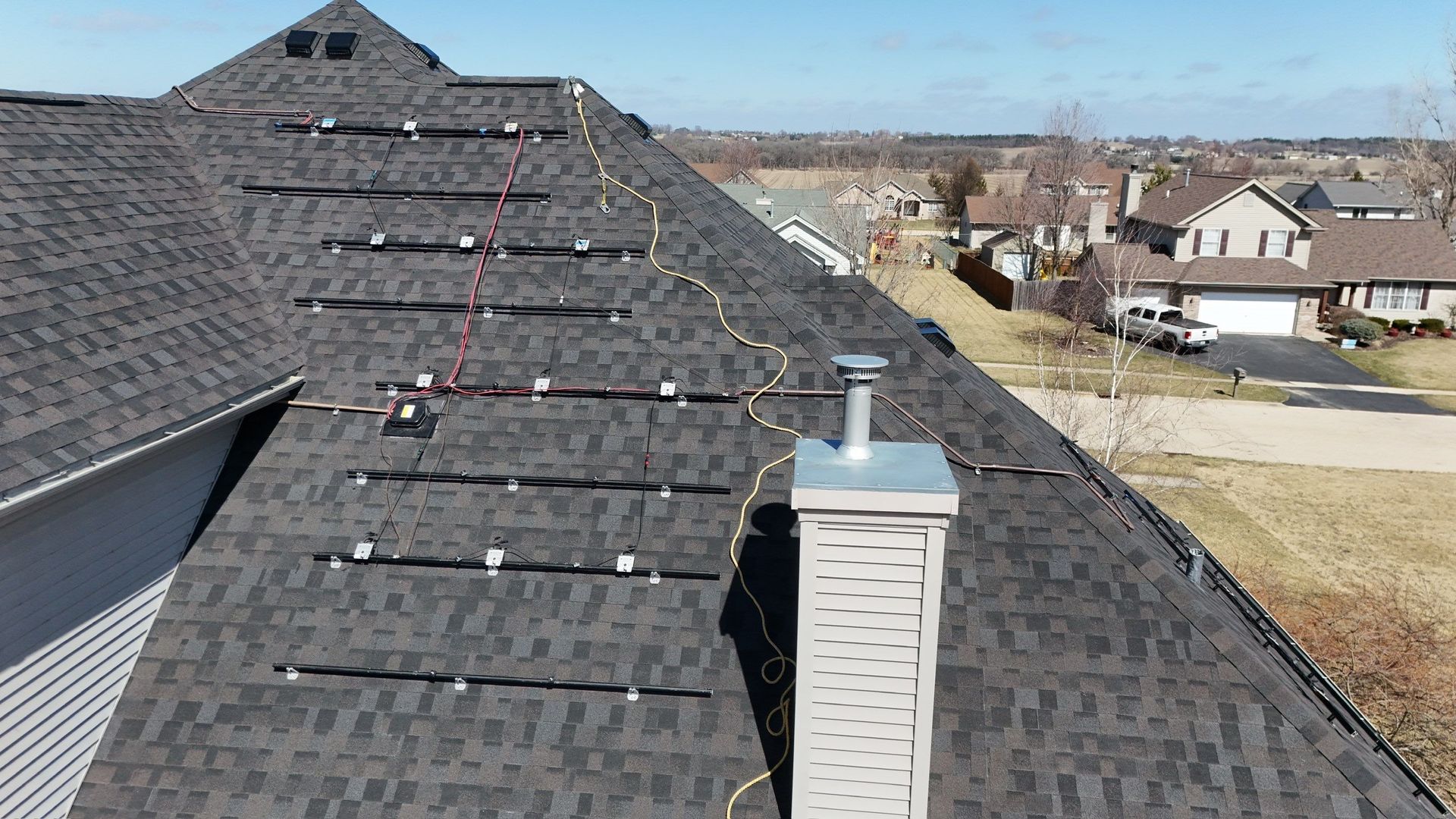 Solar panels installed on a dark-shingled roof, with a chimney and residential neighborhood in the background.