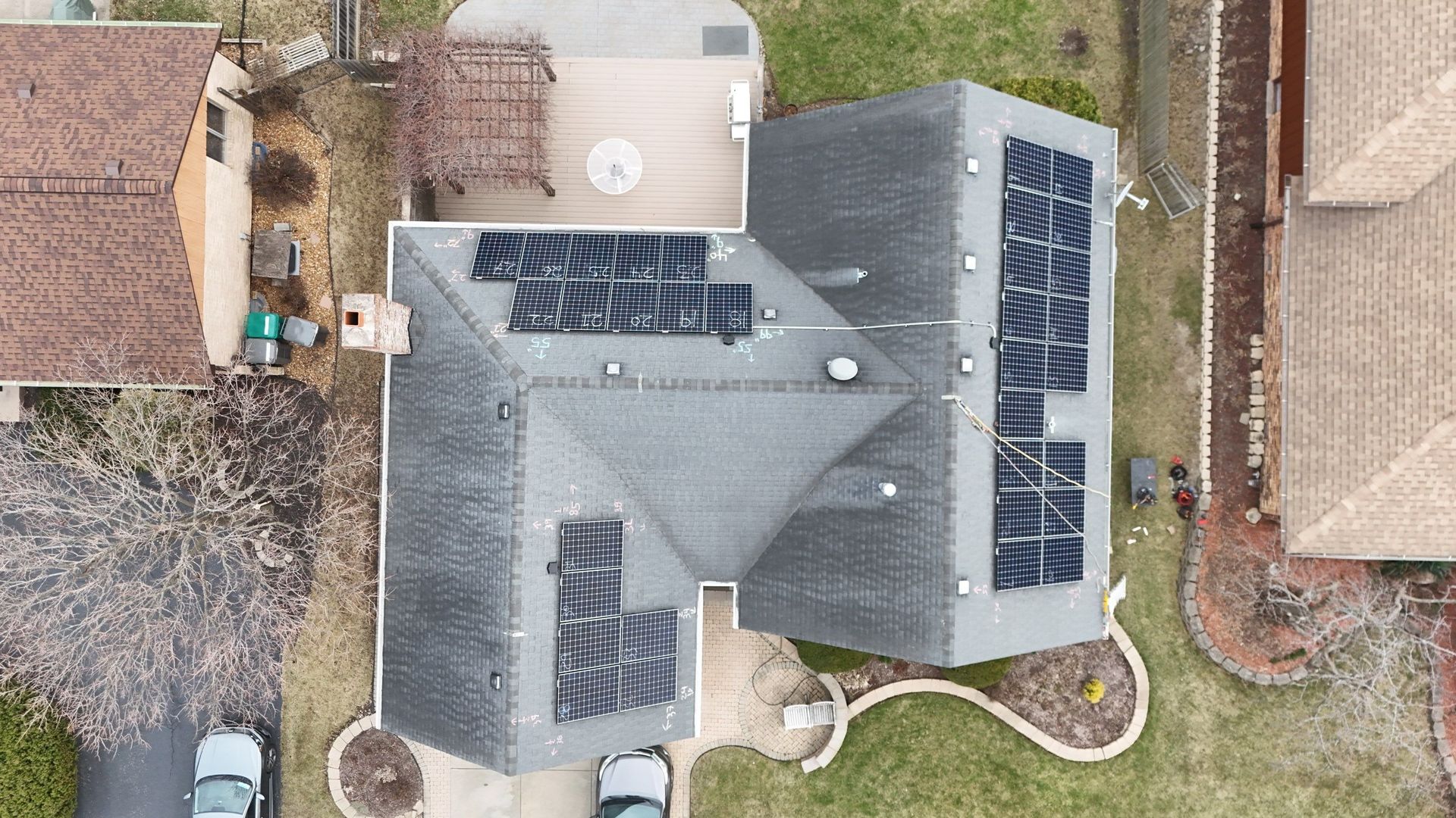 Aerial view of a house with solar panels on the roof, surrounded by grass and other houses.