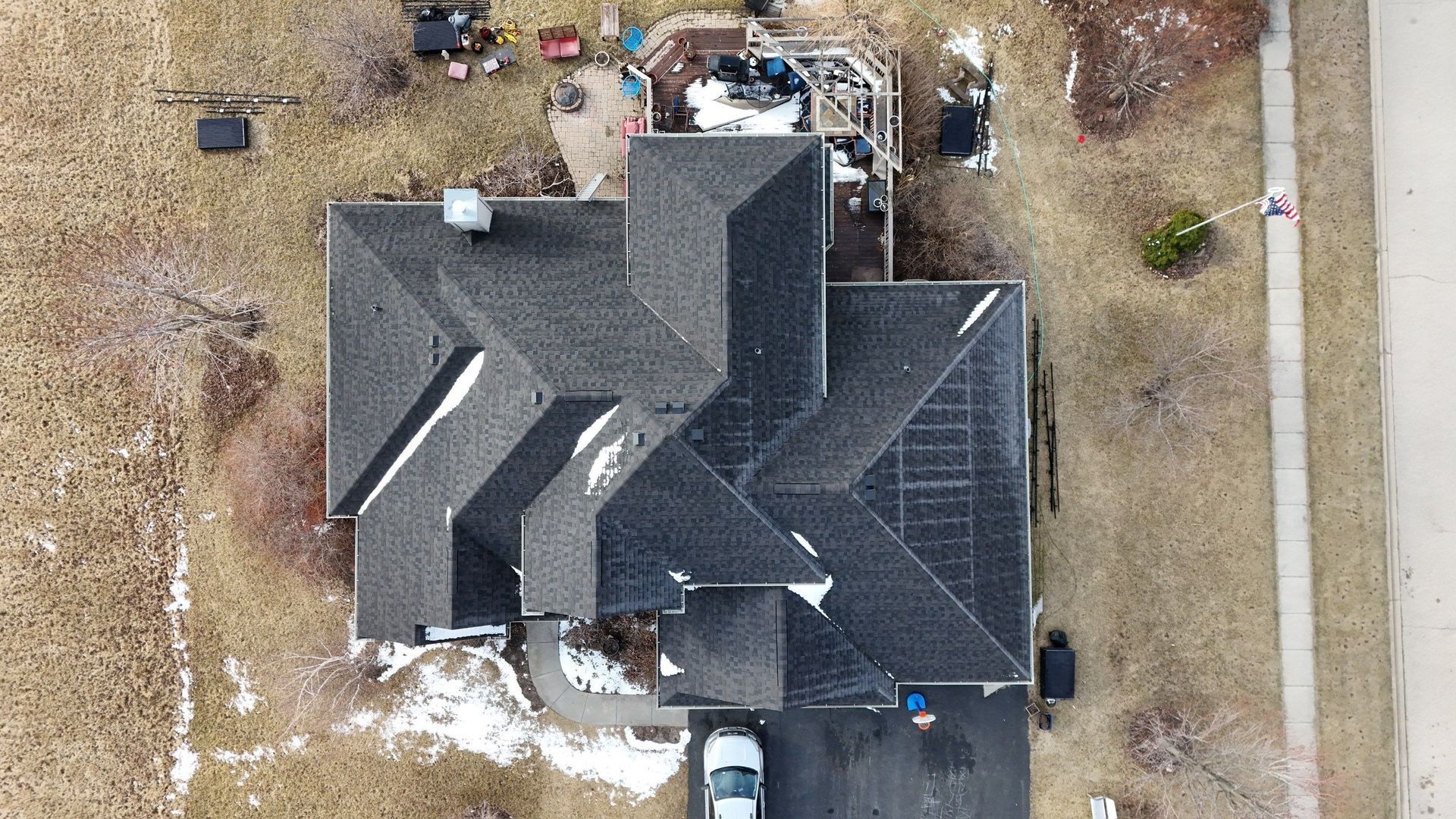Overhead view of a house with a dark roof, white car in driveway, surrounded by brown grass and a sidewalk.