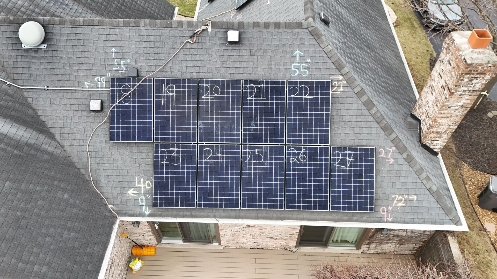 Overhead view of solar panels on a gray shingled roof; a brick chimney and yard are also visible.