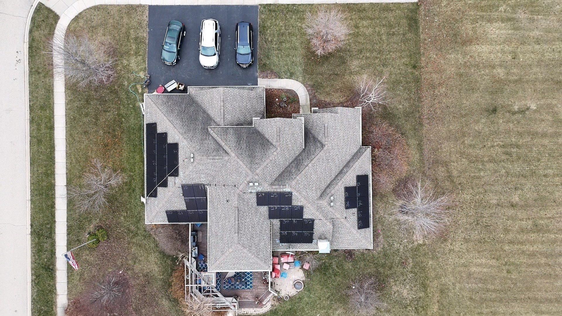 Overhead view of a house with solar panels on the roof, three cars in the driveway, and surrounded by grass and trees.