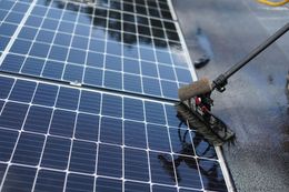 Person cleaning solar panels with a long-handled brush on a rooftop. Panels are wet and reflecting the sky.