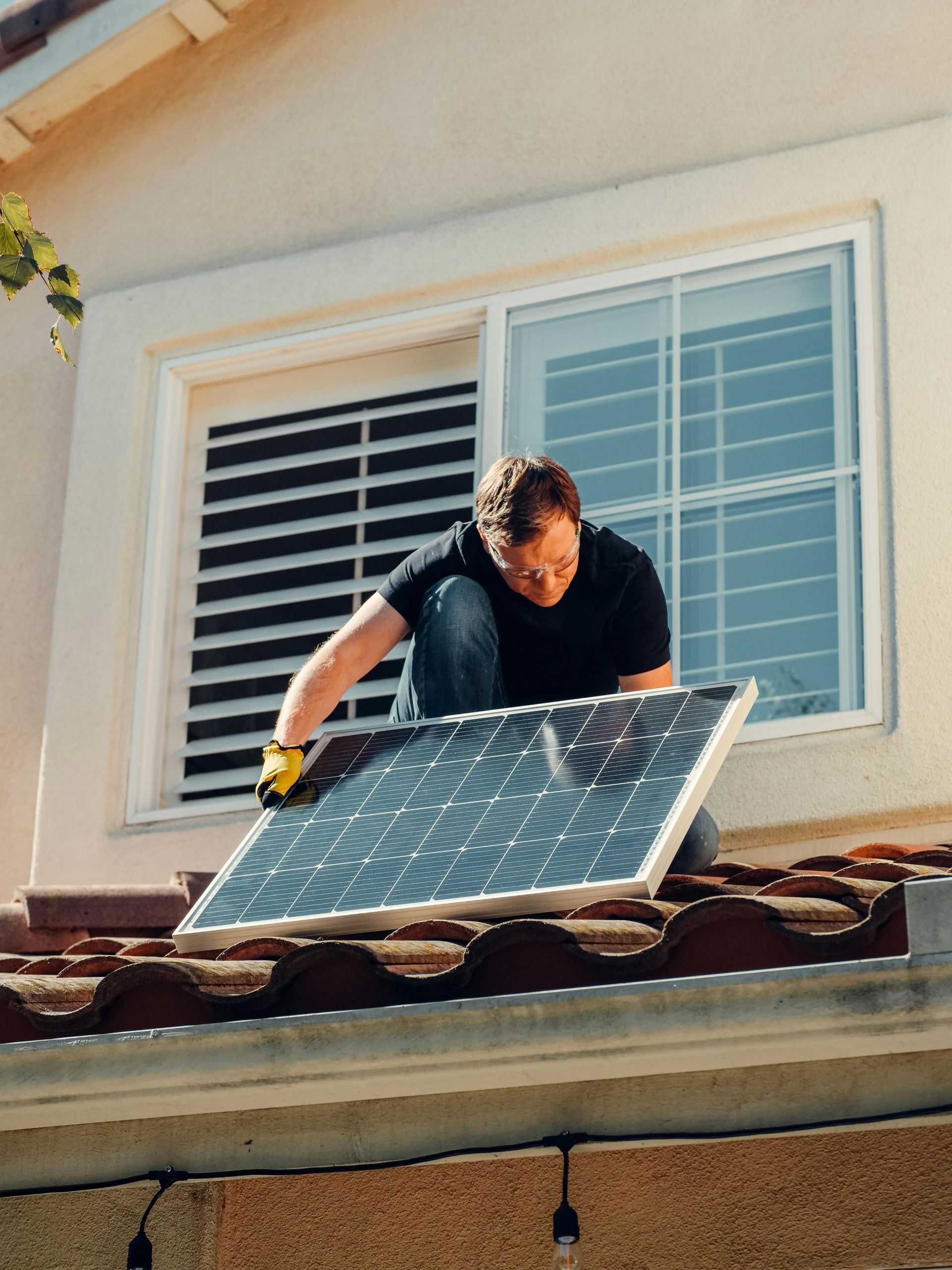 Man in black shirt installing a solar panel on a tiled roof near a window.