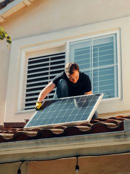 Man in black shirt installing a solar panel on a tiled roof near a window.