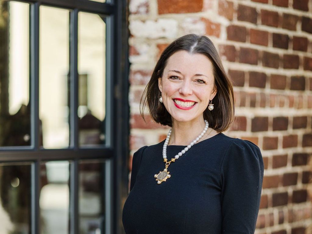 Mallory S. Morgan in a black dress and pearl necklace is smiling in front of a brick wall.