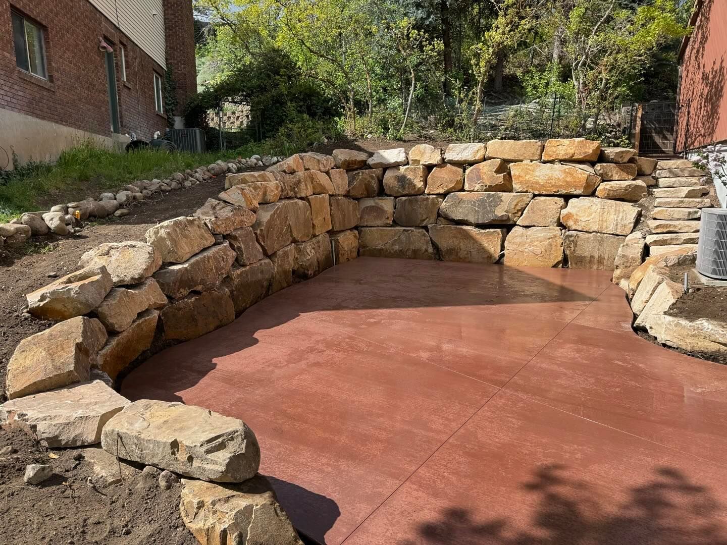 A stone wall is surrounding a concrete patio in a backyard.