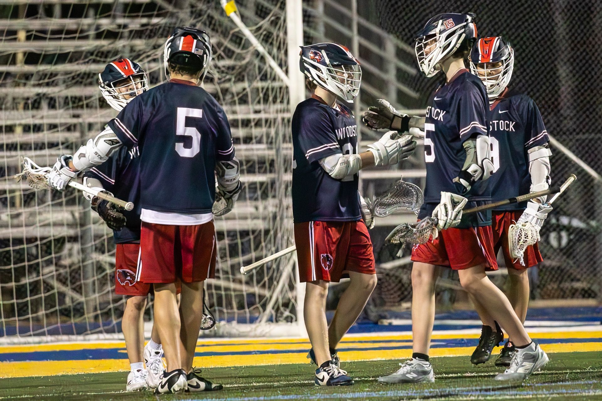 Lacrosse players in navy and red uniforms huddle on the field near the goal at night.