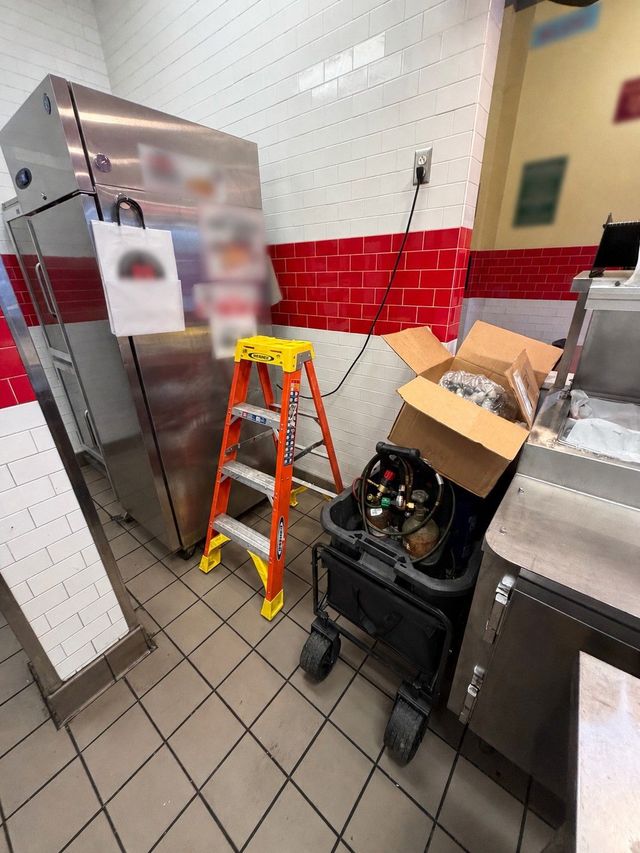 Commercial kitchen corner with ladder, stainless steel appliance, and supplies stacked by a red tiled wall.