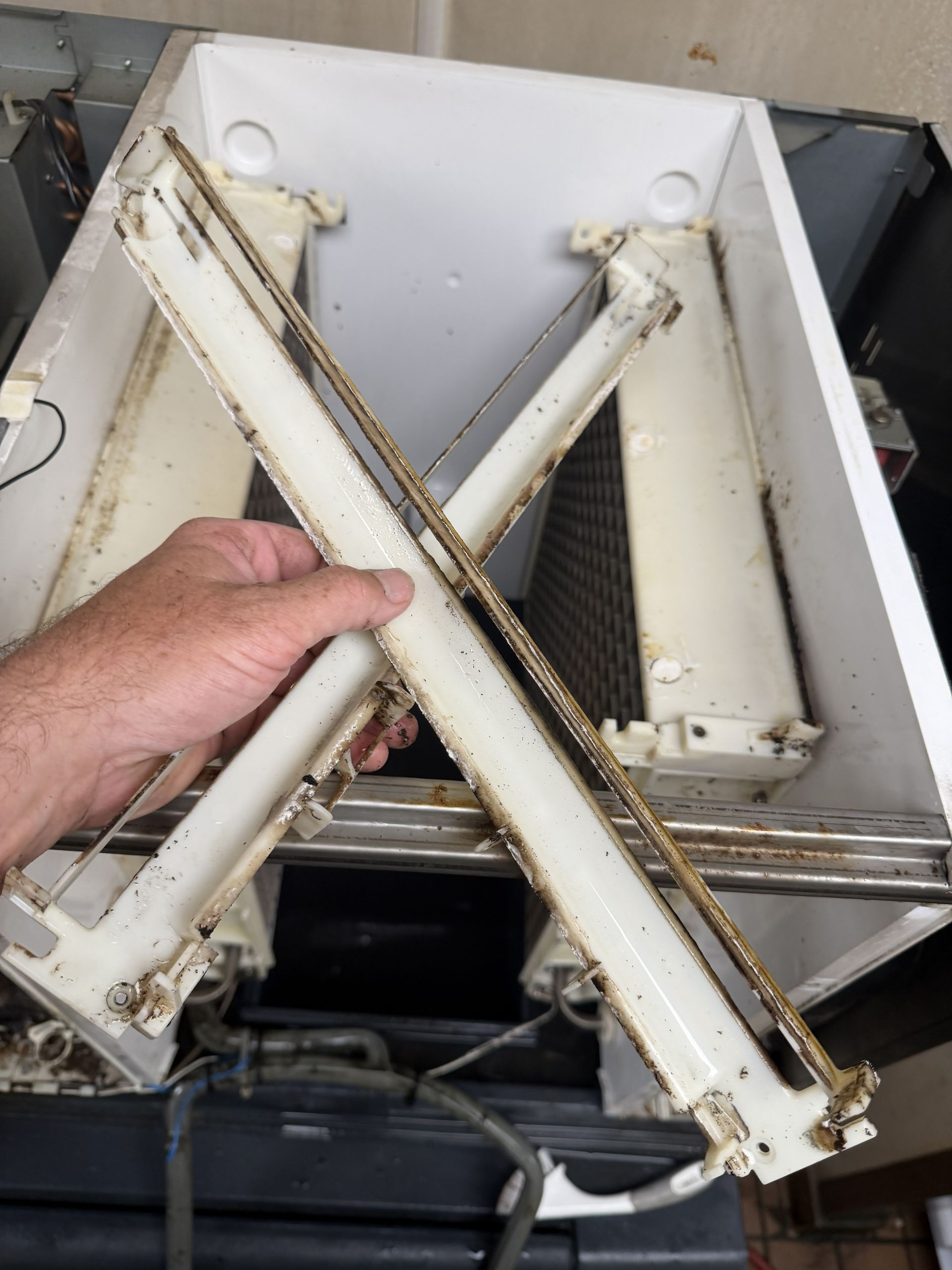 Hand holding a white metal X-shaped bracket with dirt and wear, inside a machine compartment