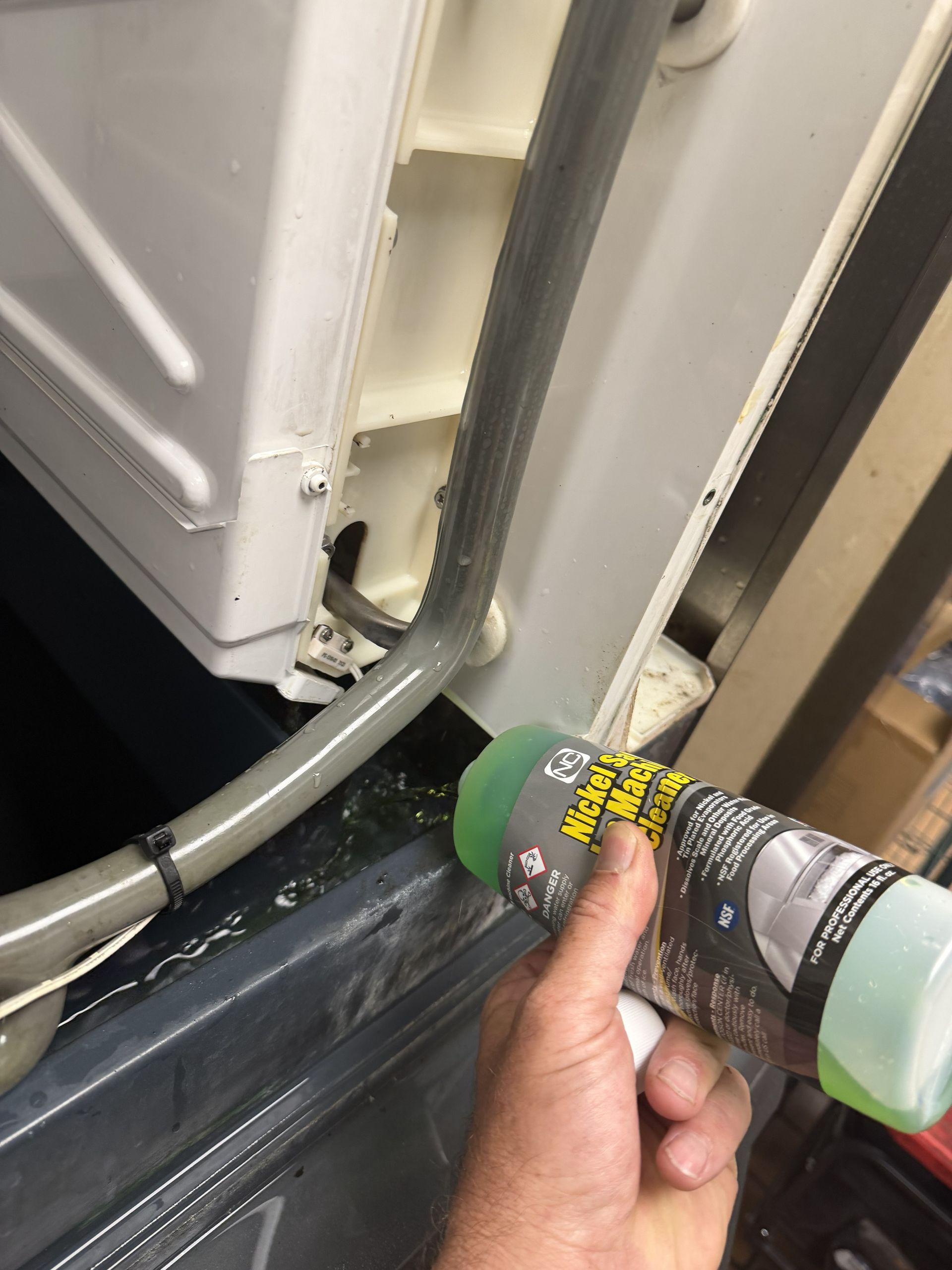 Hand using a green aerosol can to spray cleaner on a white appliance corner and nearby tubing