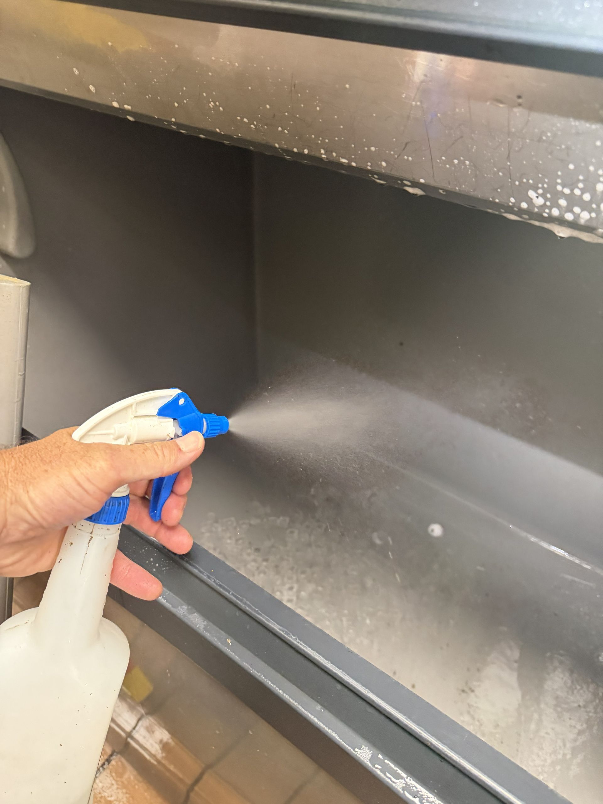 Hand spraying cleaner inside a gray metal cabinet shelf with a blue nozzle