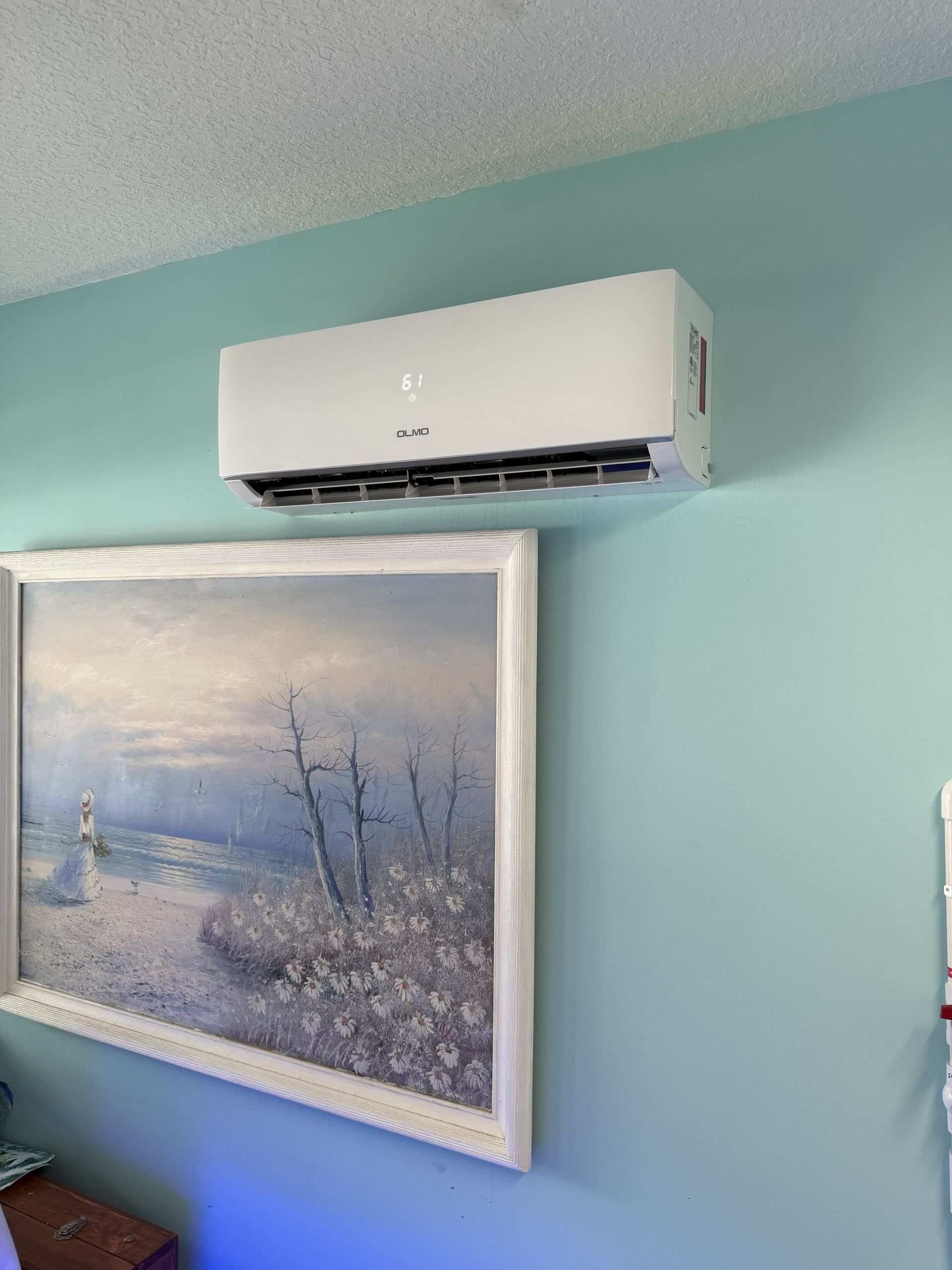 Wall-mounted white air conditioner above a framed winter landscape painting on a teal wall