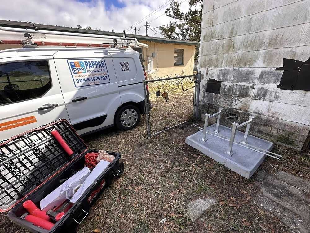White utility van beside a house, with a tool case on the ground and a metal platform under outdoor equipment.