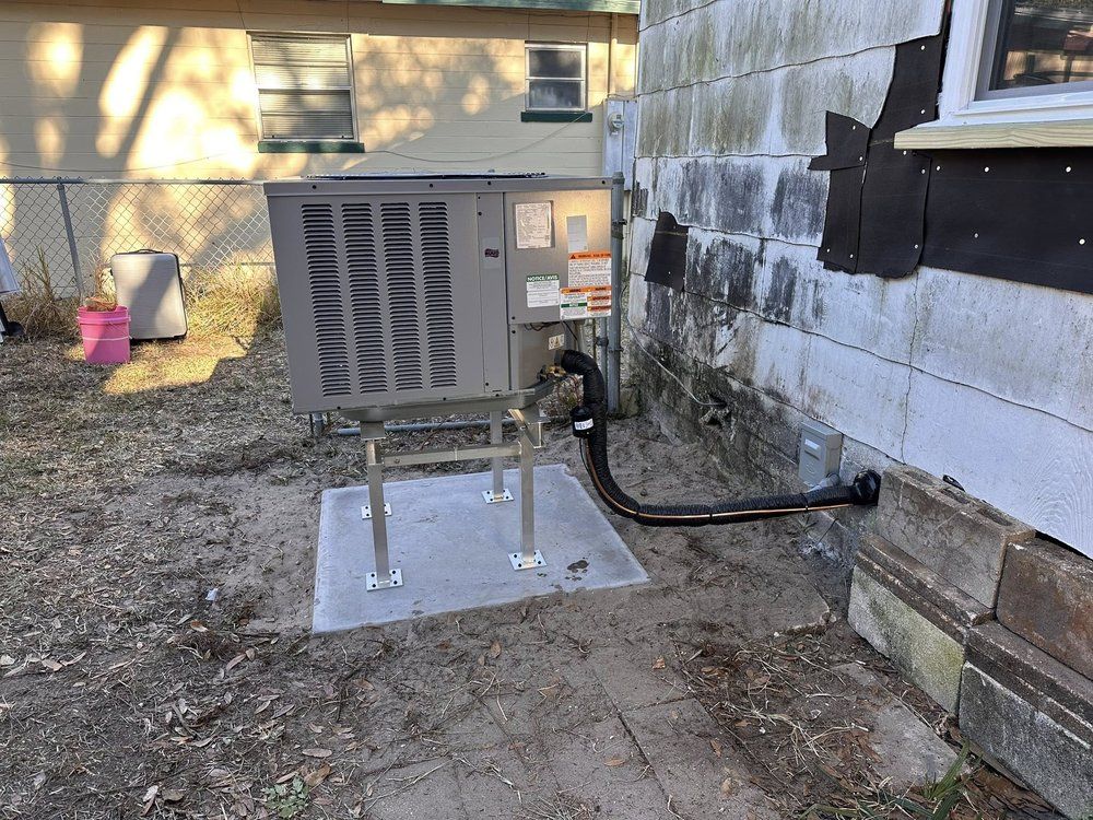 Outdoor HVAC unit beside a building, mounted on a concrete pad with black piping along the wall.