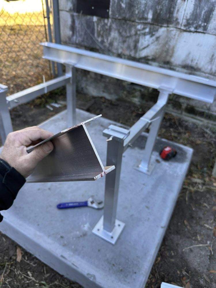 Hand holding a metal bracket near a newly mounted white steel frame on a concrete base outdoors