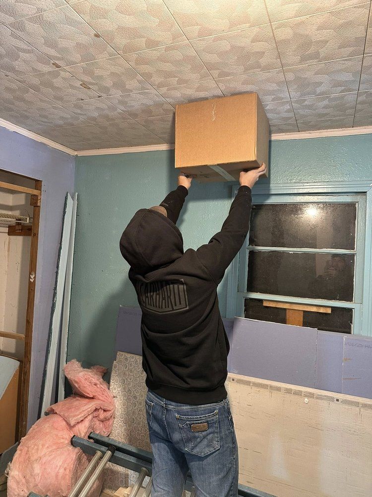 Person reaching up to lift a cardboard box toward a ceiling opening in a room.