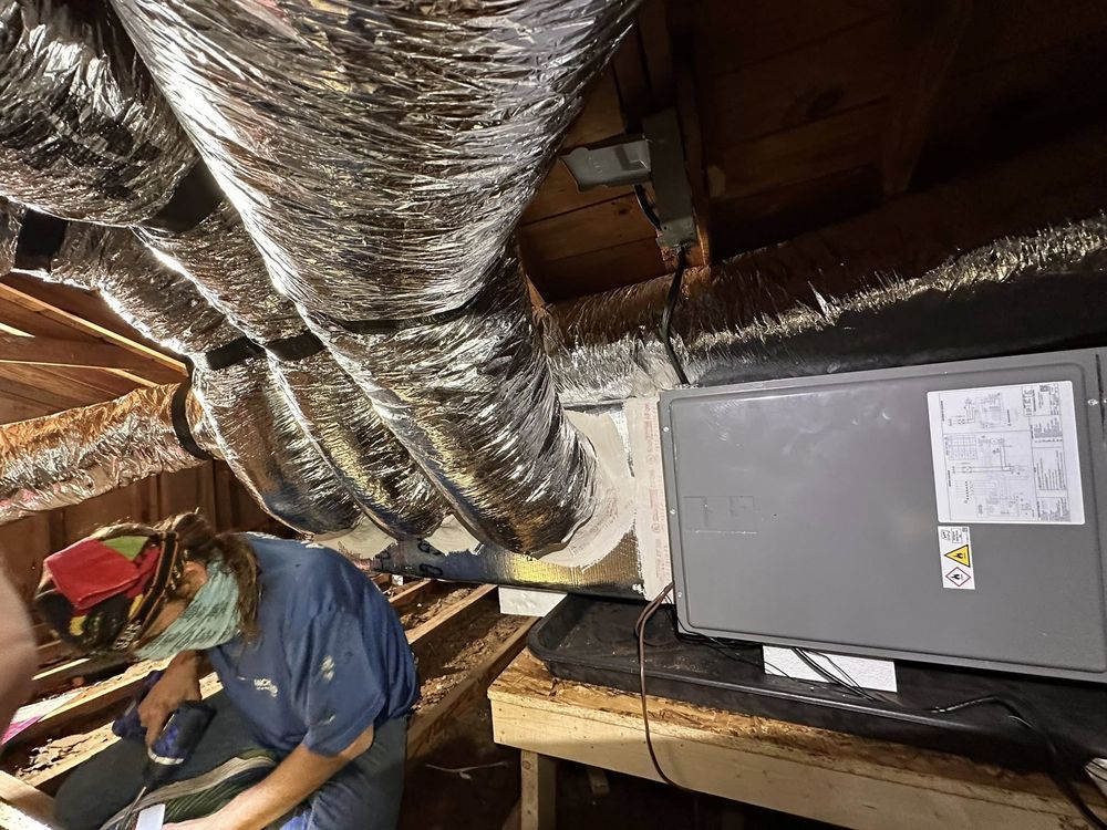 Person working in a cramped attic beside shiny ductwork and a large gray HVAC unit.